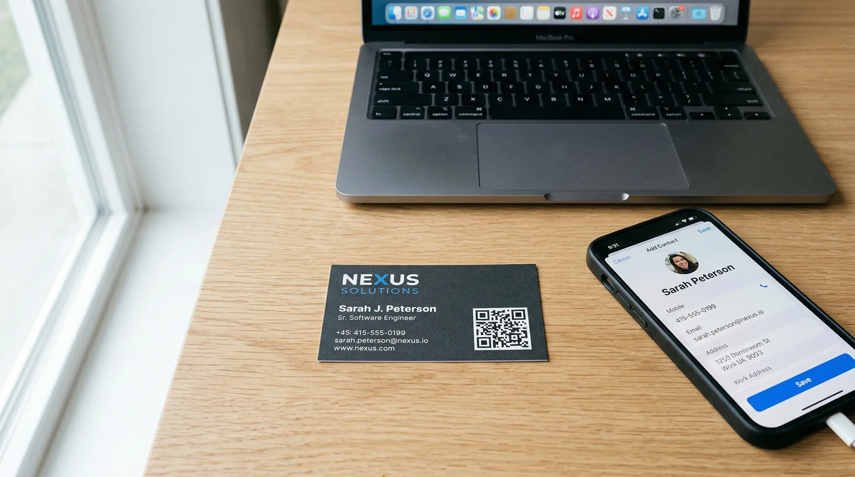 Business card with a QR code on a desk next to a phone showing an Add Contact screen with name, phone, and email filled in