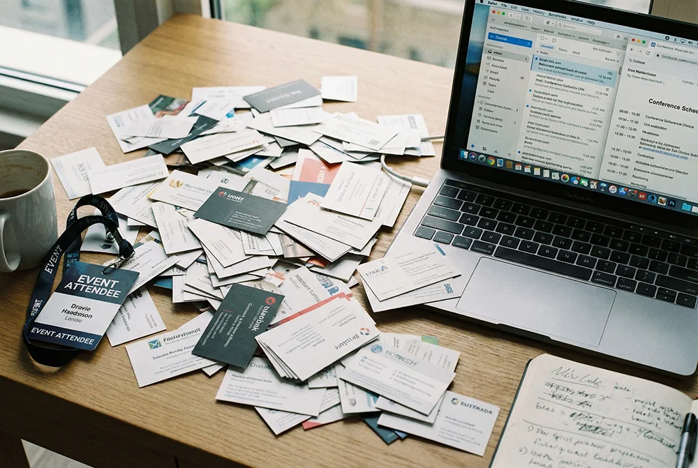 Scattered pile of paper business cards on a desk next to a laptop representing the post-networking contact chaos